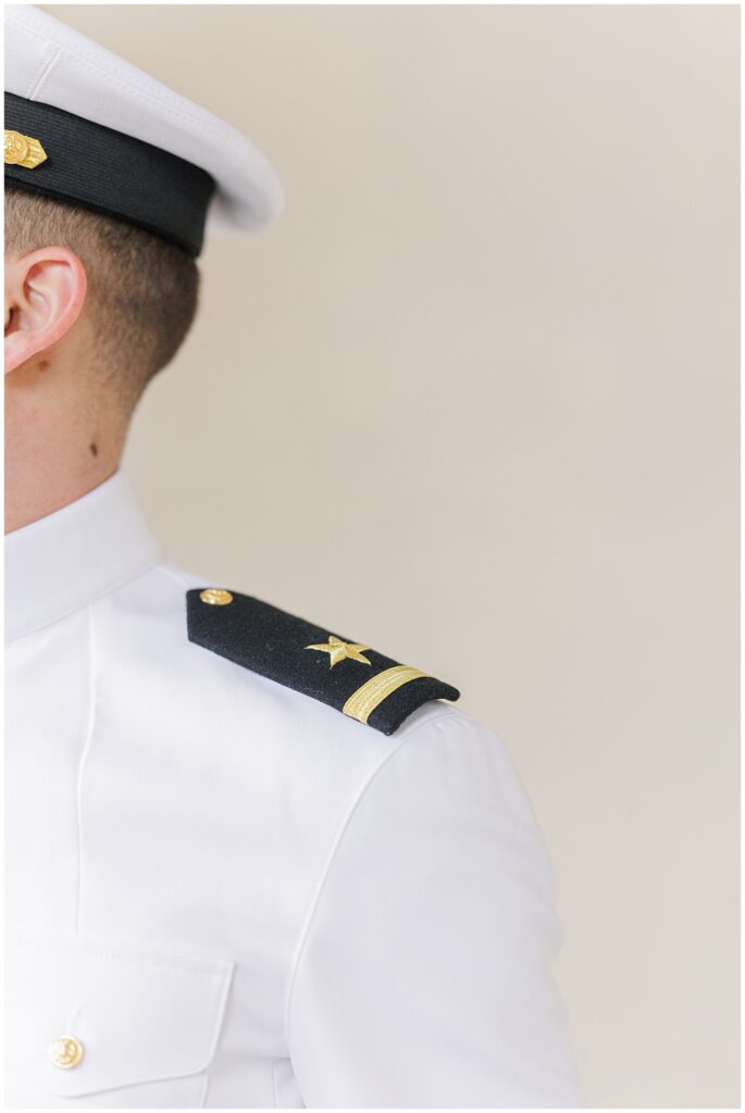 A close-up of a groom’s white Navy dress uniform showing the rank insignia on the shoulder and part of his cap, against a soft cream background.