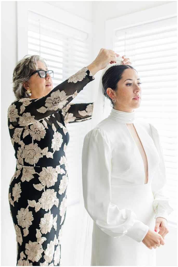 A woman in a floral black gown adjusts a veil on the bride, who wears a long-sleeved white gown with a high neckline and stands calmly in front of bright windows.
