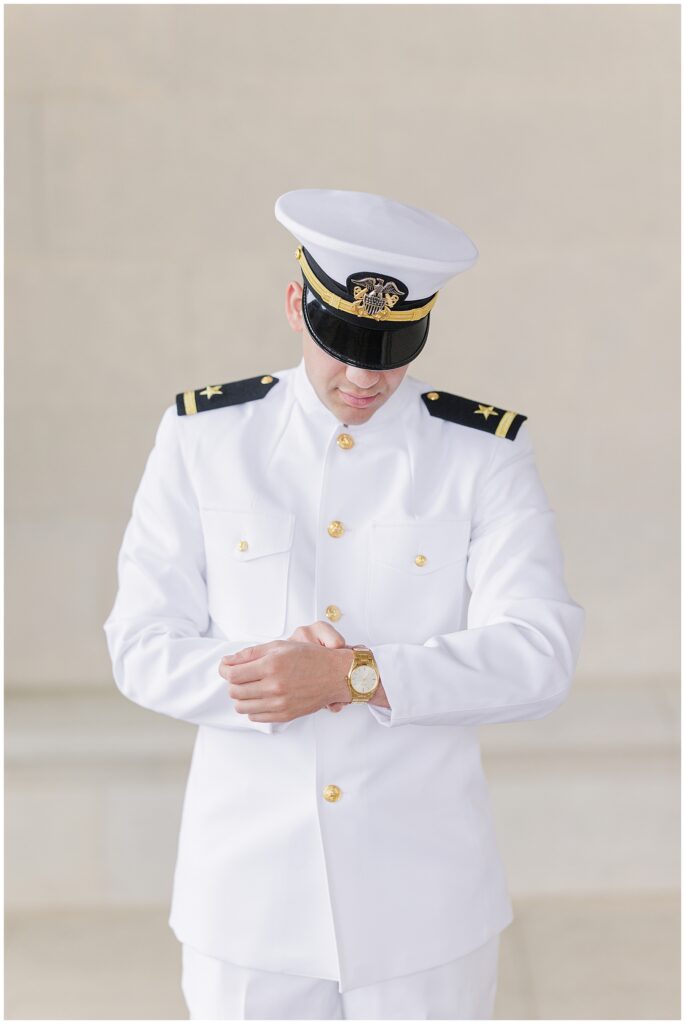 A groom in a U.S. Navy dress white uniform looks down while adjusting his gold wristwatch, captured during a Washington D.C. wedding.