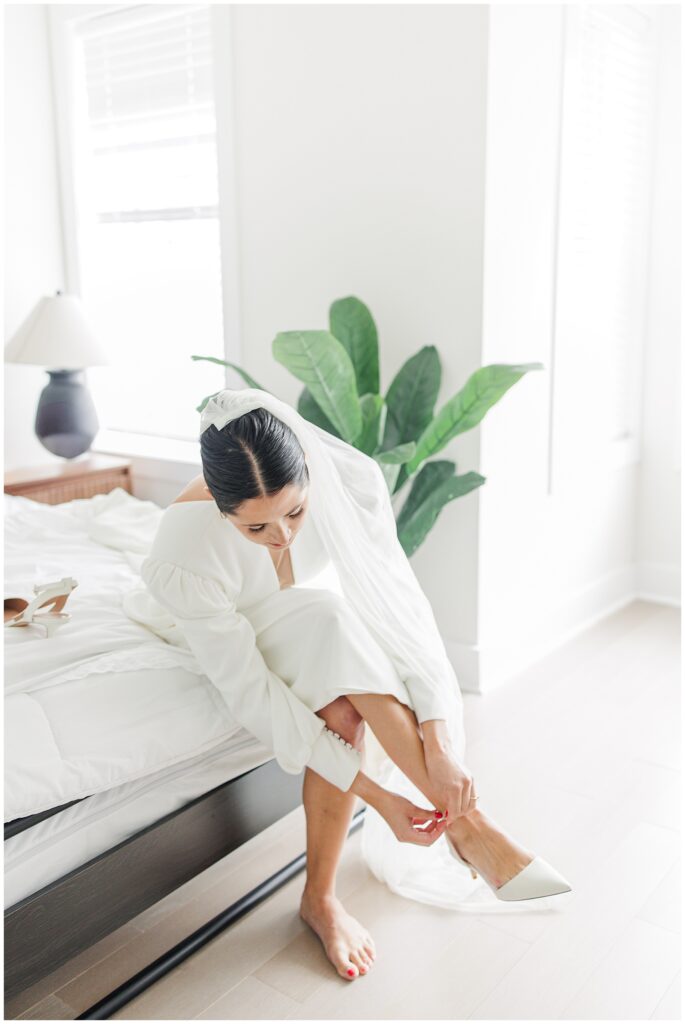 The bride, seated on the edge of a bed in a white room, bends forward to strap on one white heel, her veil draped behind her.