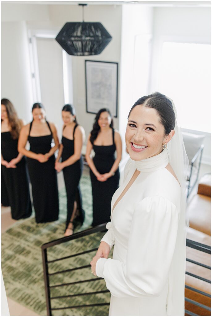 A smiling bride in a white gown and veil stands at the top of a staircase while her bridesmaids, dressed in black, wait below.