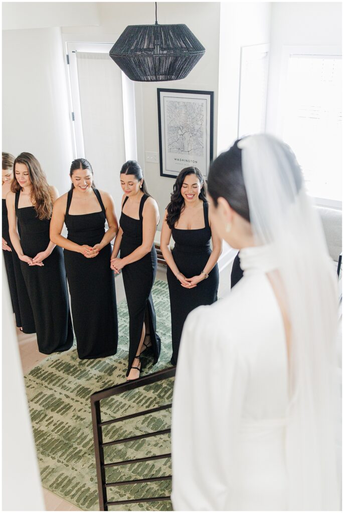 A view over the bride’s shoulder as her bridesmaids, all in matching black gowns, react with smiles and admiration in a well-lit room.