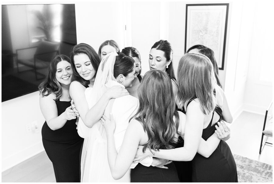 Black-and-white photo of a bride in a white gown and veil being embraced in a tight group hug by her bridesmaids dressed in black gowns, captured by a Washington D.C. wedding photographer.