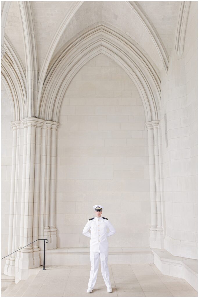 A groom in a white military dress uniform stands under the tall arched stone architecture of the Washington National Cathedral, hands behind his back, looking forward.