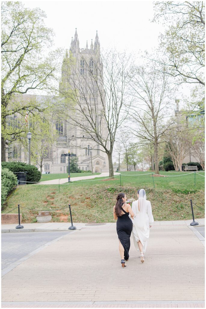 A bride in a white gown and veil walks hand-in-hand with a bridesmaid in a black dress along a path outside the Washington National Cathedral, with the cathedral’s Gothic tower visible in the background.