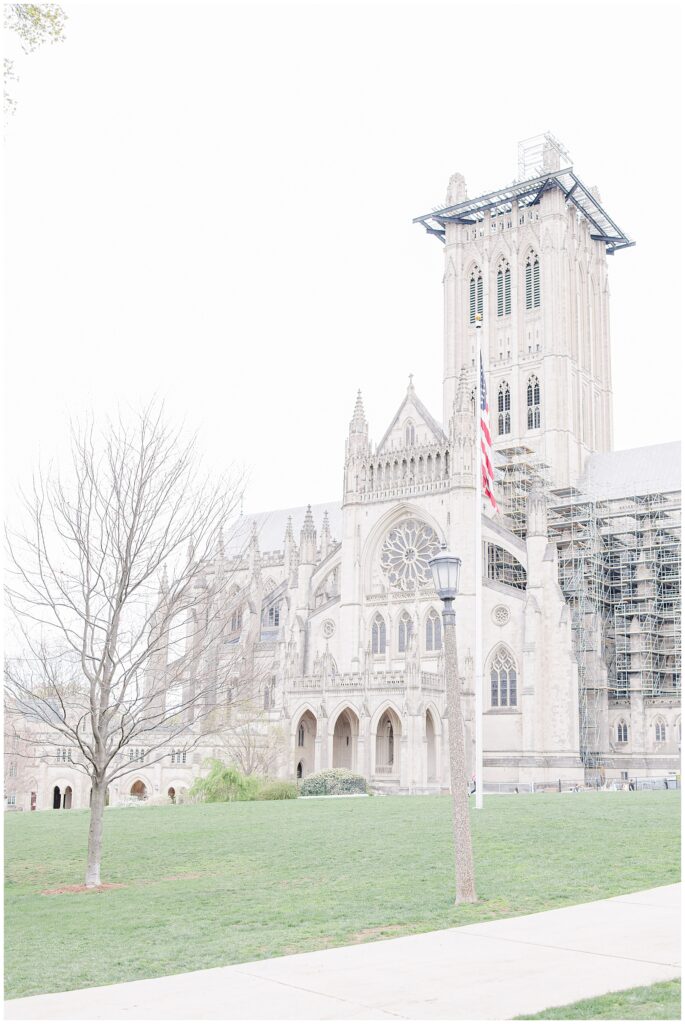 A wide view of the Washington National Cathedral’s exterior shows its Gothic architecture, tall tower with scaffolding, and an American flag on a pole in the foreground.
