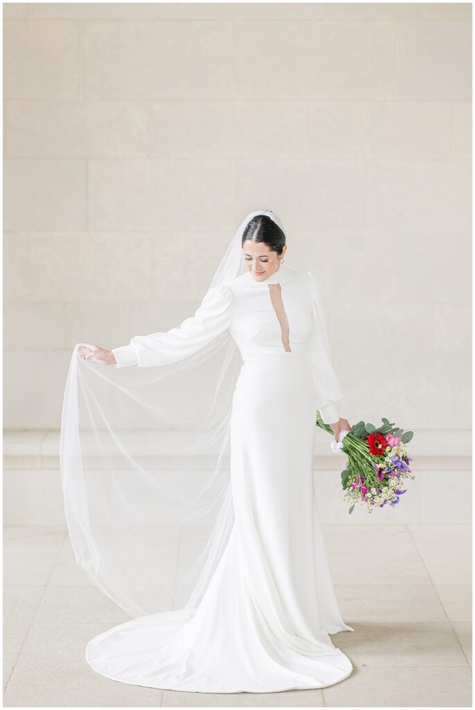 A bride in a fitted long-sleeve gown and veil holds her bouquet in one hand and lifts part of her veil with the other, standing under the arches of the Washington National Cathedral.