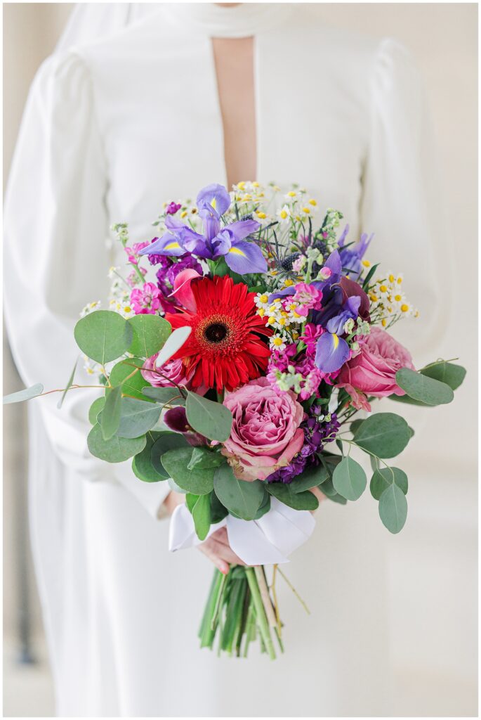 Close-up of a colorful bridal bouquet with red, purple, pink, and white flowers held against a white gown during a Washington National Cathedral wedding.