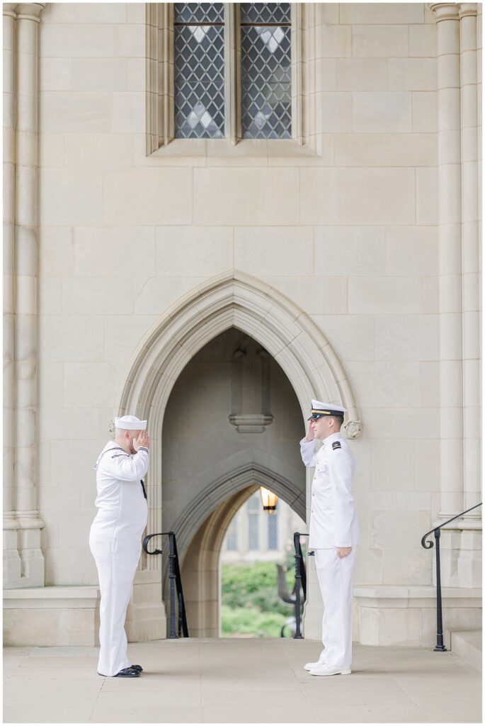 Two men in white military uniforms face each other and exchange a salute in front of a stone arched entryway at the Washington National Cathedral.