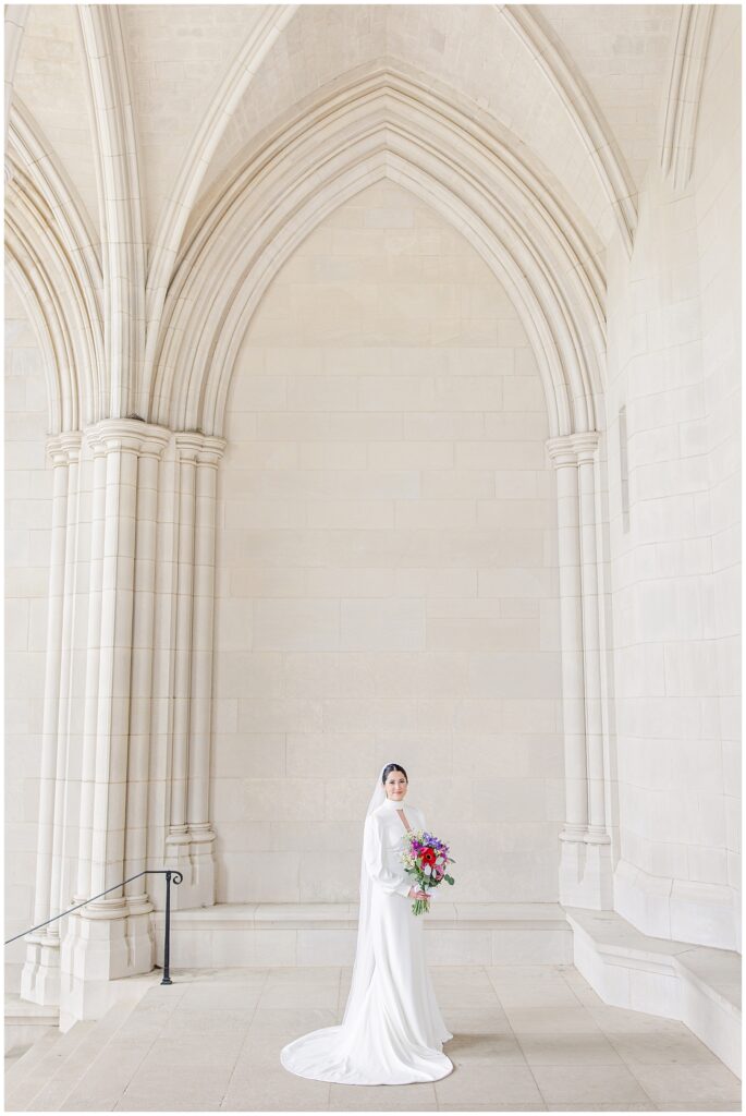 A bride in a long-sleeve gown and veil stands centered beneath the soaring stone arches inside the Washington National Cathedral, holding a colorful bouquet.