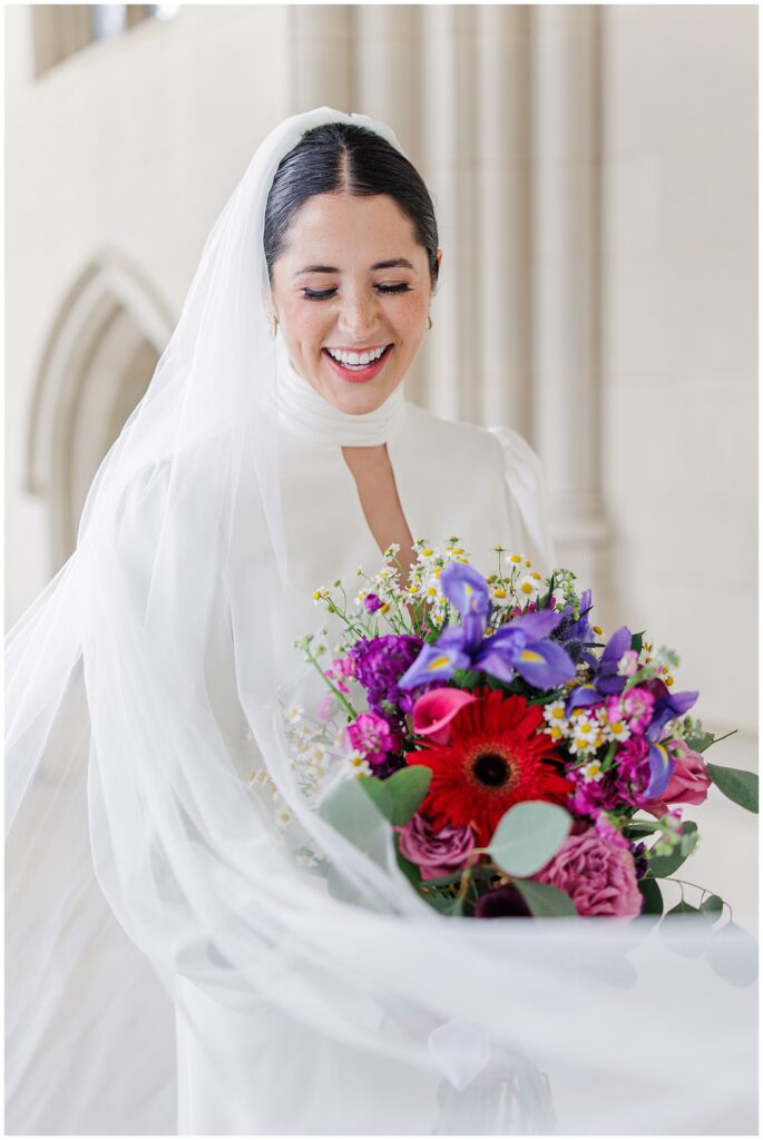 A smiling bride in a white gown and veil holds a vibrant bouquet of colorful flowers during her Washington National Cathedral wedding.