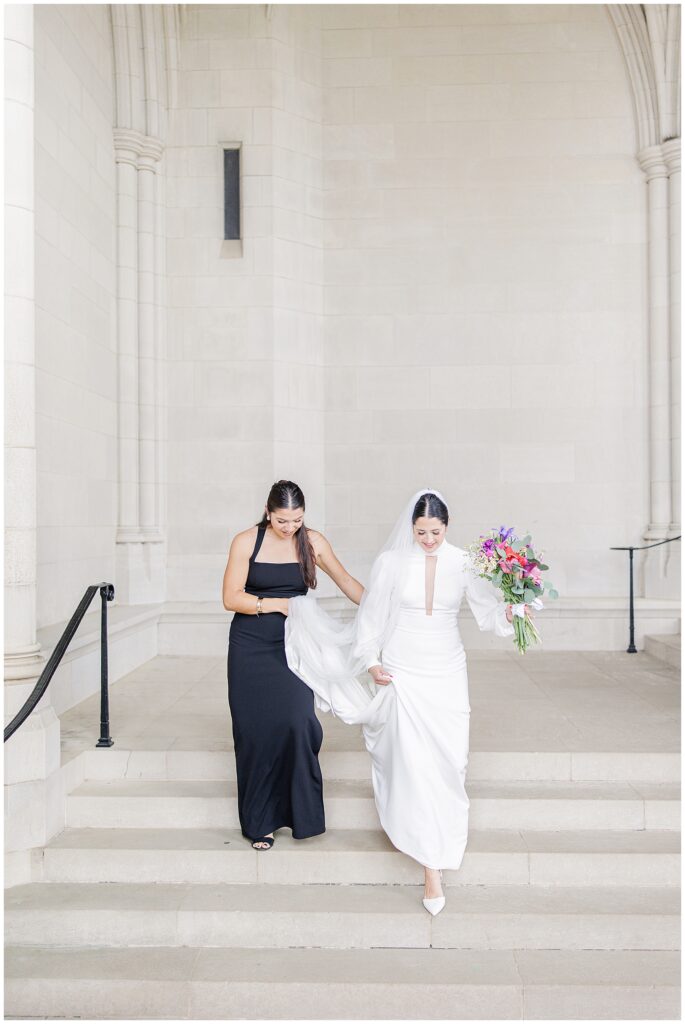 A bride and a bridesmaid walk down stone steps inside the Washington National Cathedral, the bride holding her gown and bouquet while the bridesmaid assists with her train.