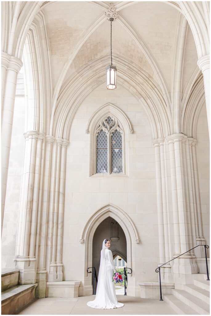 A bride stands centered beneath a tall arched doorway and ornate stone columns at the Washington National Cathedral, facing the camera with her bouquet in hand.