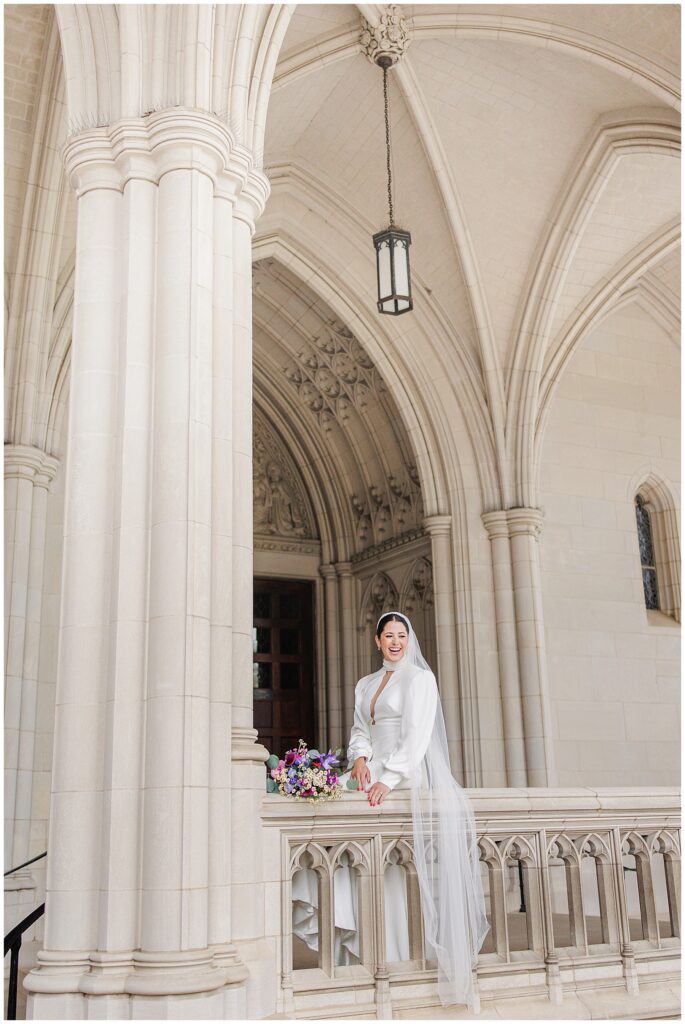 A bride leans on a stone balcony ledge, laughing, with her bouquet beside her, surrounded by the grand arches of the Washington National Cathedral.