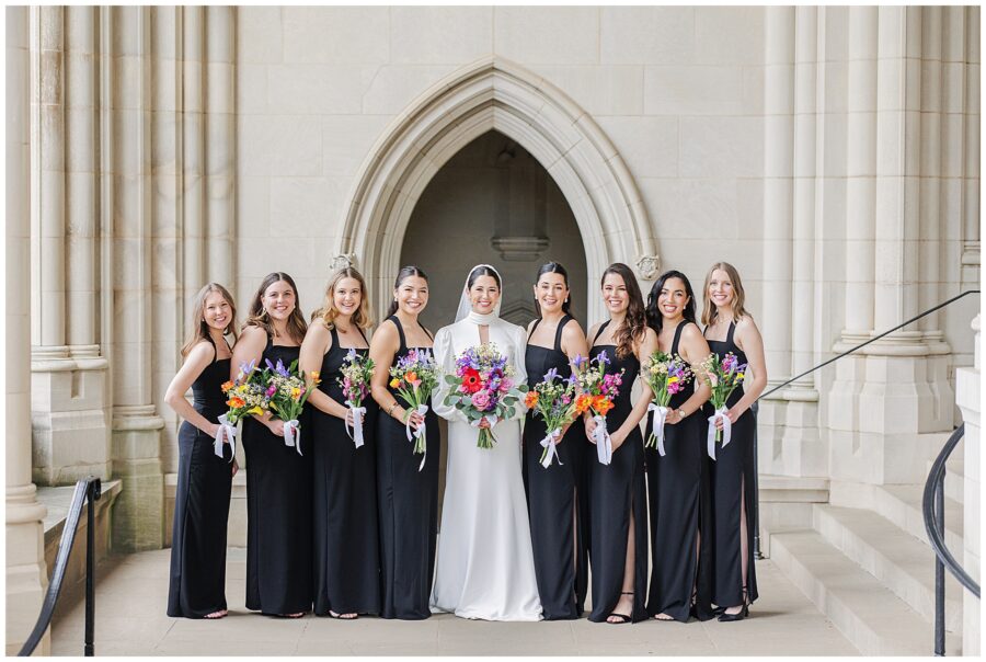 A bride stands with her bridesmaids in black dresses, each holding a colorful bouquet, in front of an arched stone entryway at the Washington National Cathedral.
