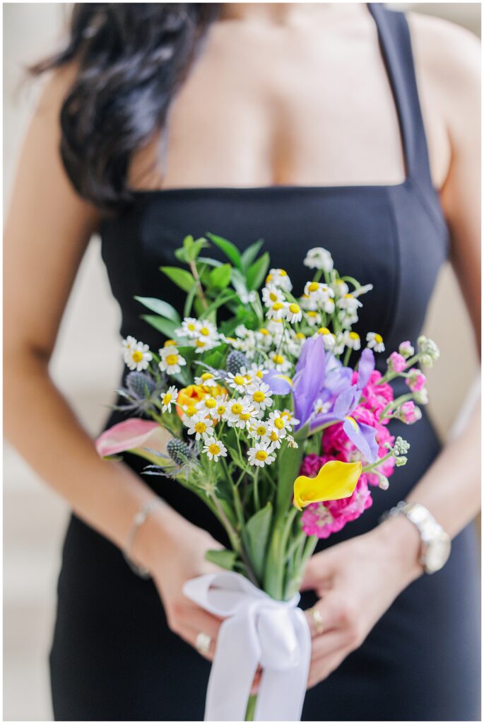 A bridesmaid in a black dress holds a bouquet of wildflowers, including daisies, yellow calla lilies, and purple irises, tied with a white ribbon, during a Washington National Cathedral wedding.