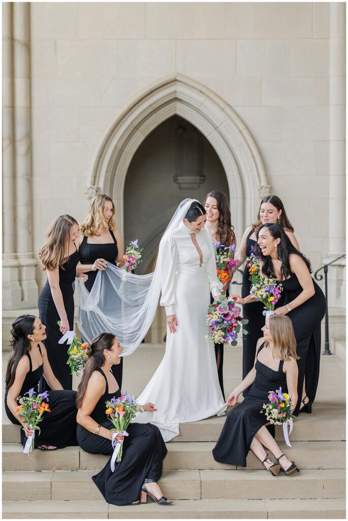 A bride surrounded by bridesmaids adjusts her veil on the stone steps of the Washington National Cathedral, all smiling and holding colorful bouquets.