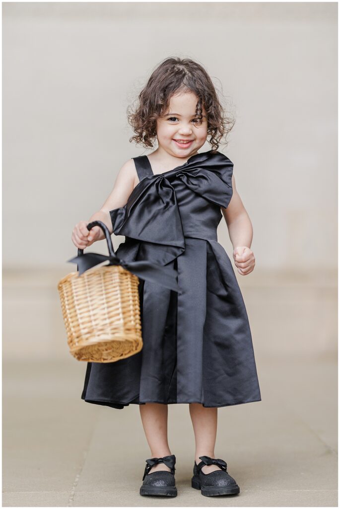 A young flower girl in a black satin dress with a large bow holds a woven basket and smiles during a Washington National Cathedral wedding.