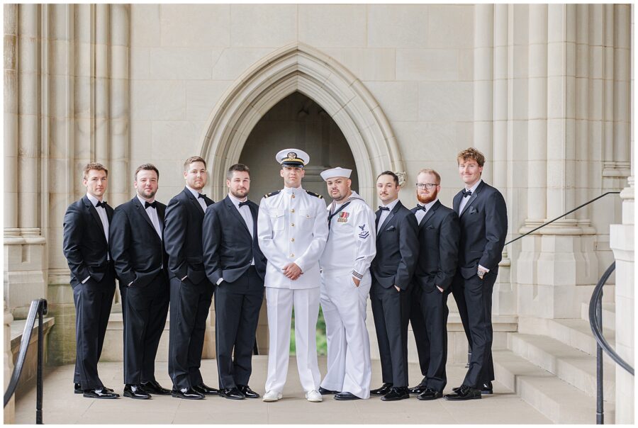 The groom in a white military dress uniform stands with his groomsmen in black tuxedos and another man in a Navy uniform in front of a stone archway at the Washington National Cathedral.