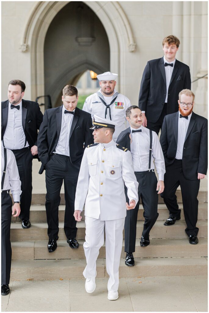 The groom in a white military dress uniform walks down the steps with his groomsmen in black tuxedos and another man in a Navy uniform in front of a stone archway at the Washington National Cathedral.
