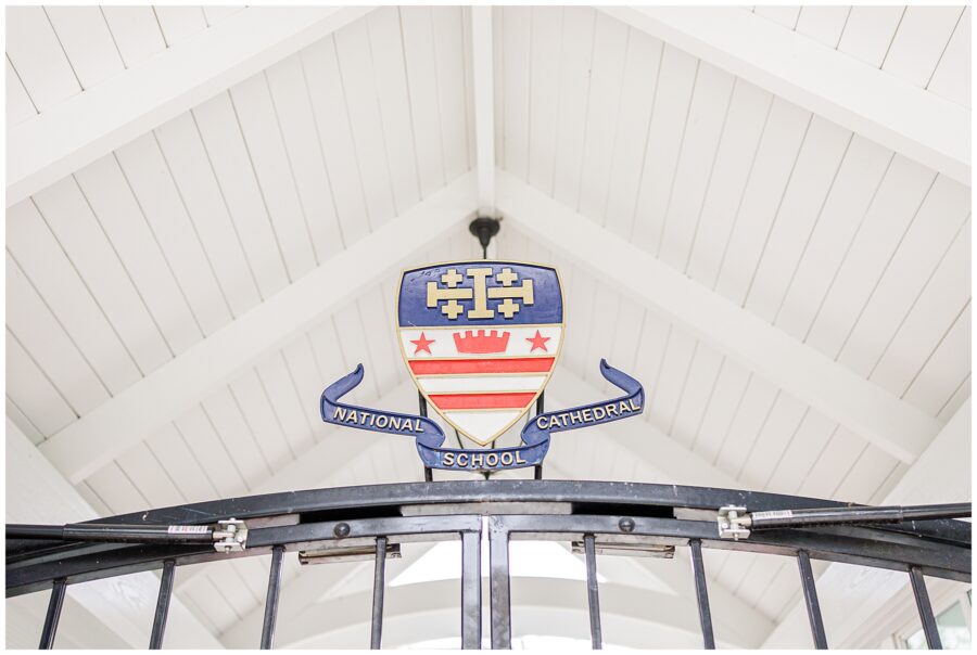 Close-up of the National Cathedral School crest mounted on a black gate, under a peaked white ceiling.