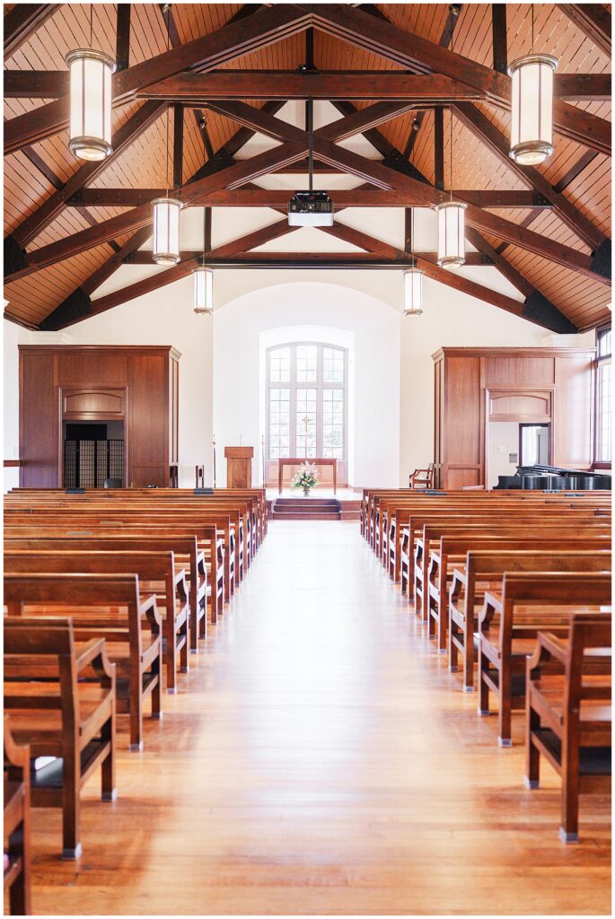Vertical view of the National Cathedral School chapel interior, focusing on the exposed wood beams and ceiling lights above the central aisle and altar.