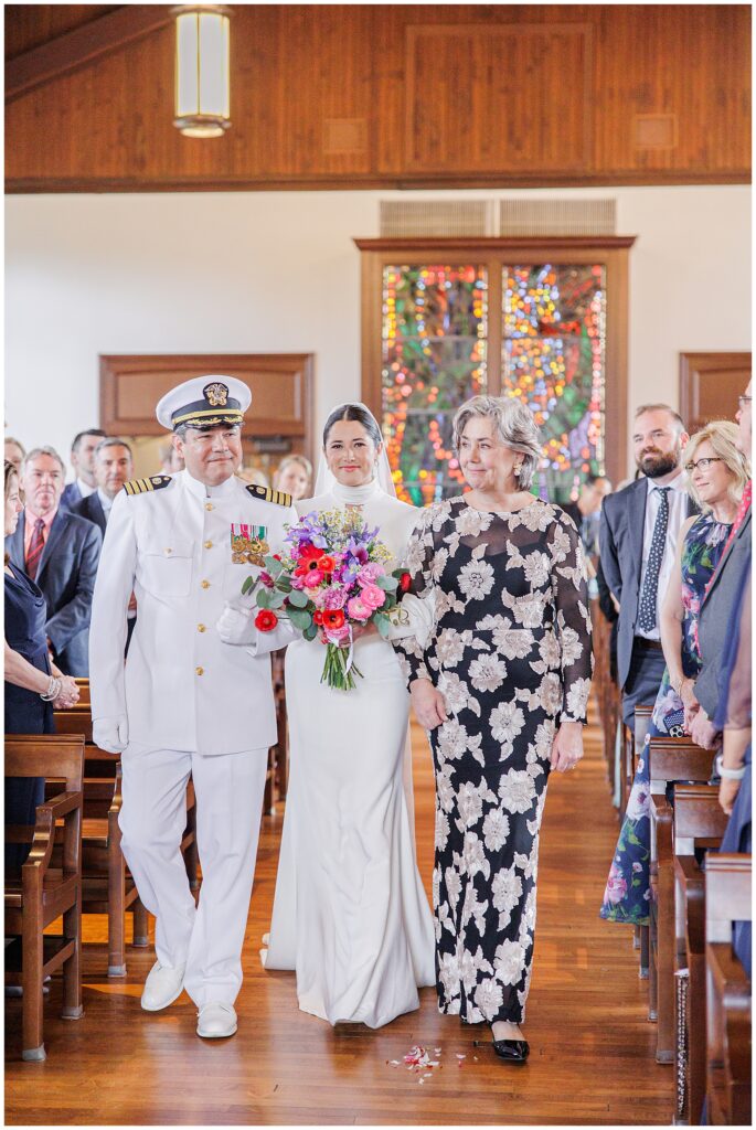 A bride in a white gown walks down the aisle at the National Cathedral School chapel, escorted by a man in a white military uniform and a woman in a black floral dress, with guests standing on either side.