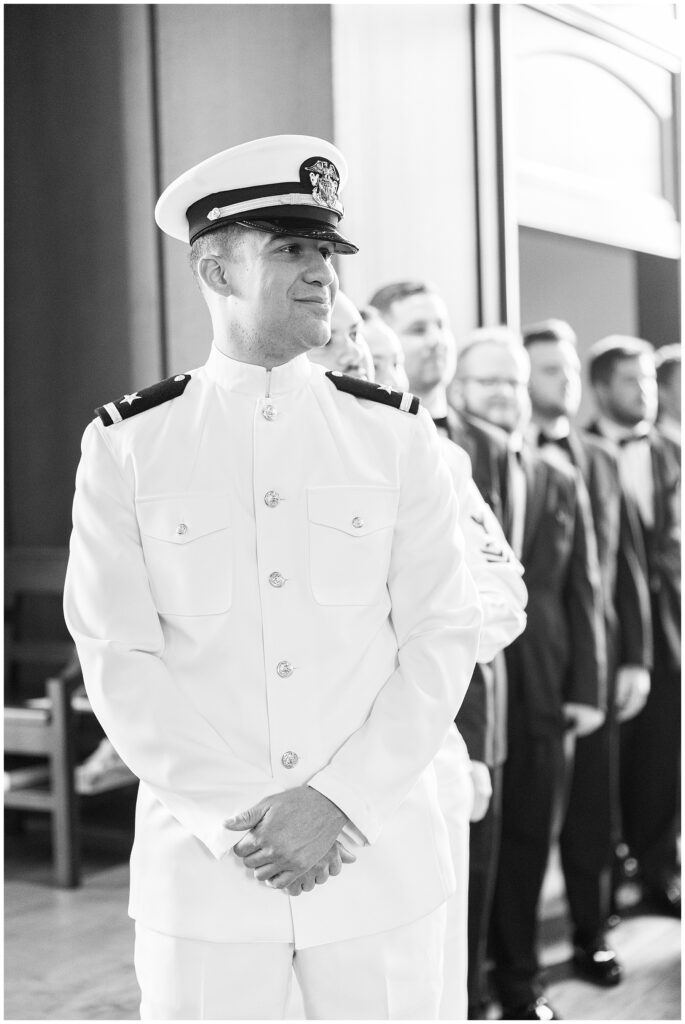 A groom in a white military dress uniform smiles and stands at the front of the chapel with groomsmen behind him during the National Cathedral School wedding ceremony.