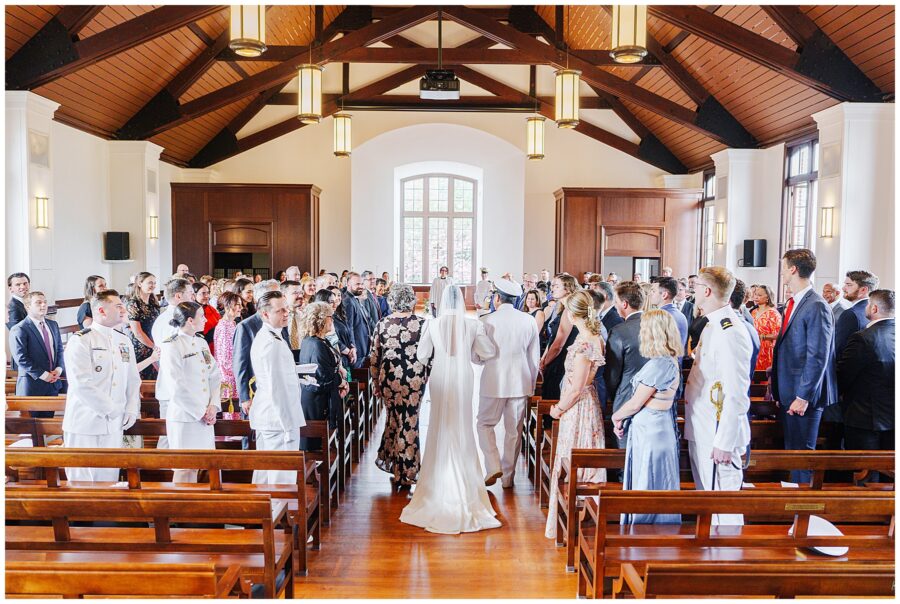Wide view of guests standing as the bride walks down the center aisle toward the altar in the National Cathedral School chapel.