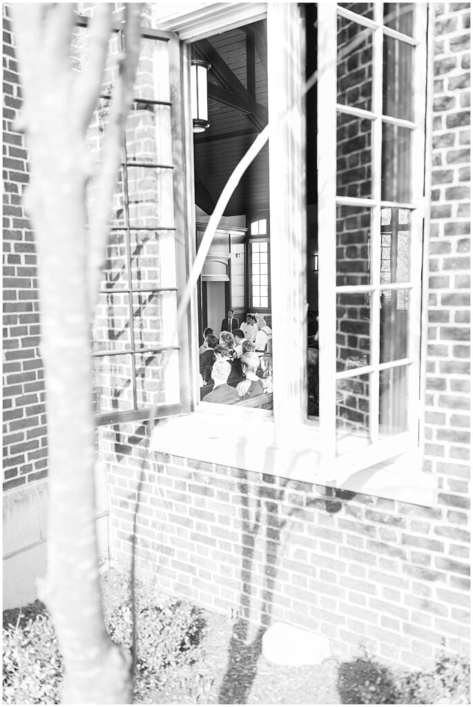 Black-and-white photo taken from outside a brick building, looking through an open window at seated guests and wedding activity inside the National Cathedral School chapel.
