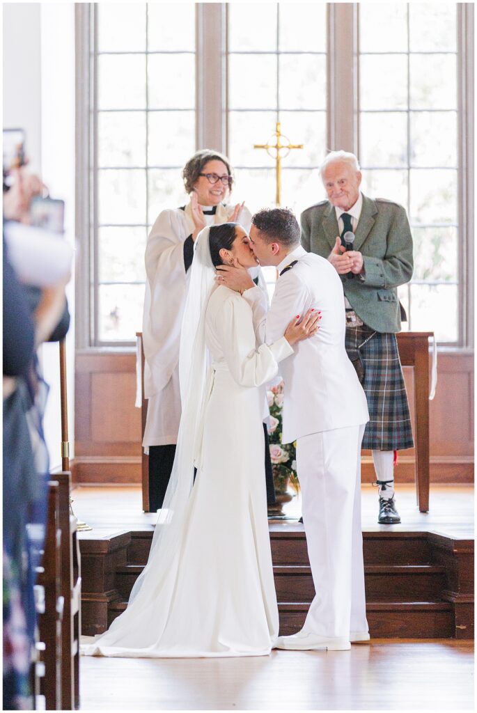 The bride and groom share their first kiss at the altar during a wedding ceremony at the National Cathedral School chapel, with two officiants smiling and clapping behind them.