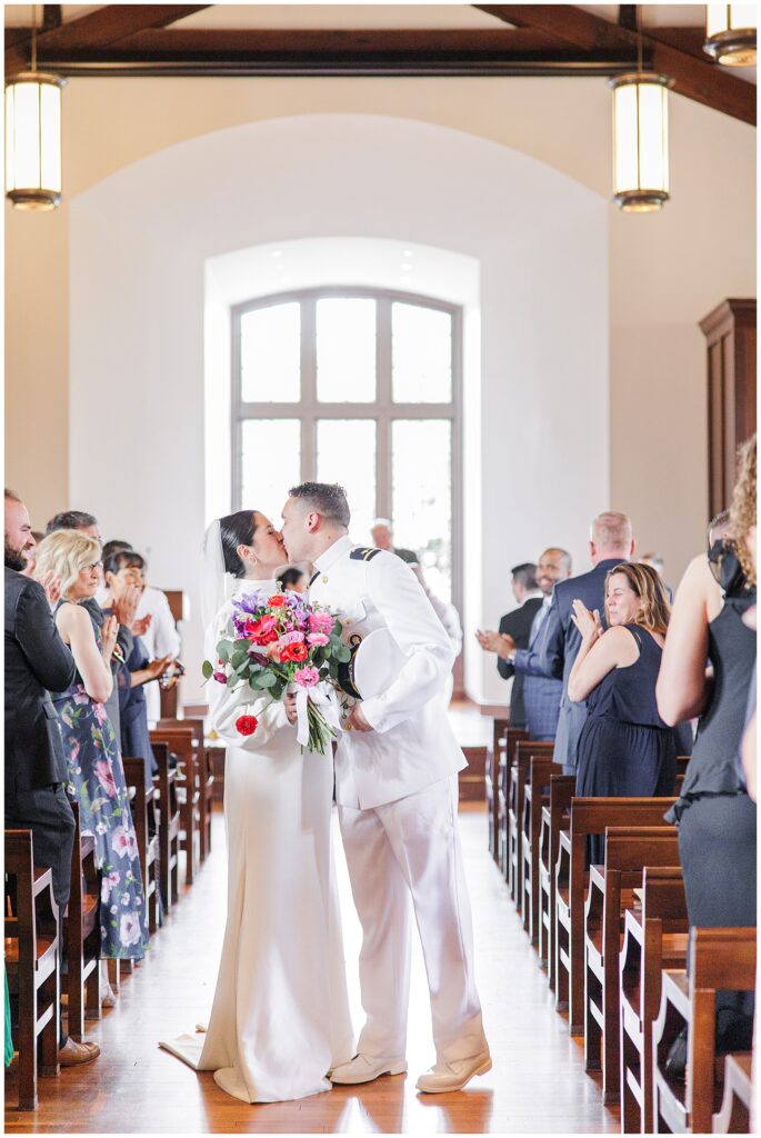 The newly married couple kisses at the end of the aisle, surrounded by applauding guests inside the National Cathedral School chapel.
