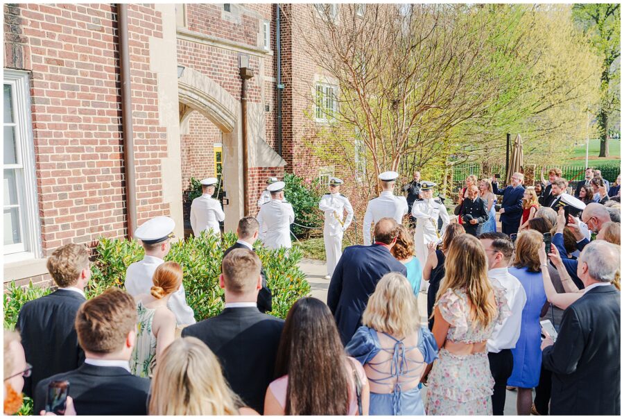 Guests gather outside a brick building at the National Cathedral School as uniformed military officers form an arch with ceremonial swords for the couple’s exit.