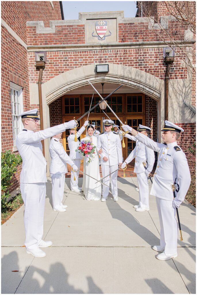 The bride and groom stand smiling beneath a traditional military sword arch outside the National Cathedral School, surrounded by officers in white dress uniforms.