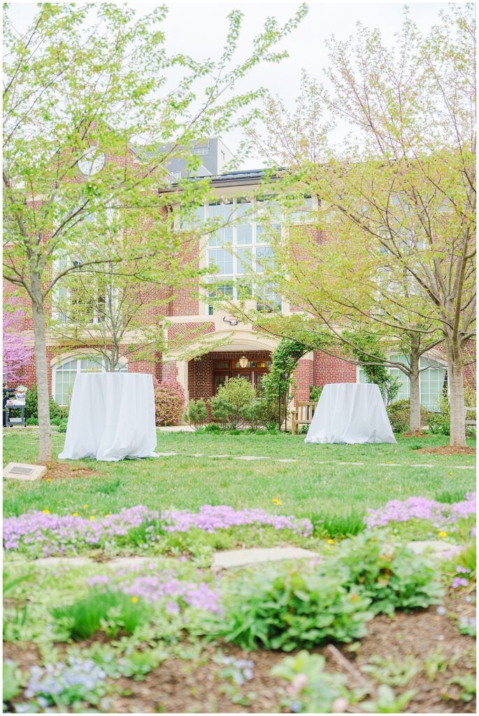 A cocktail hour setup at the National Cathedral School, with cocktail tables covered in white linens placed on a lawn surrounded by blooming trees and a red brick building.