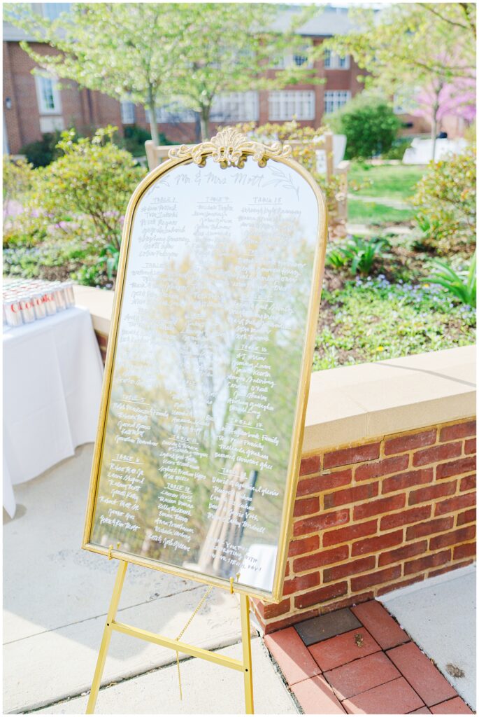 A large mirror with a gold frame and handwritten seating chart reflects the outdoor surroundings during cocktail hour at the National Cathedral School.