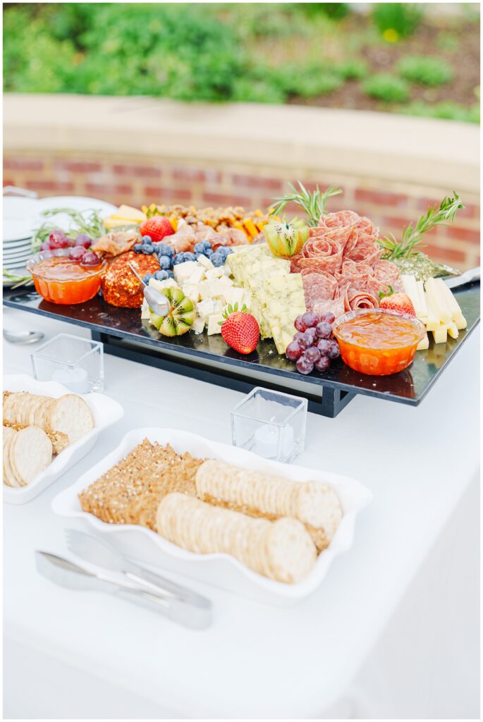 A charcuterie board on a white tablecloth at the wedding cocktail hour, featuring assorted cheeses, meats, fruits, and dips, with trays of crackers and small candles nearby.