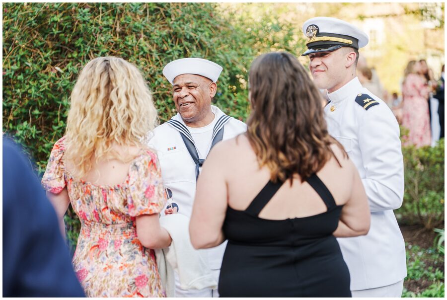 A small group of wedding guests chatting outdoors during the cocktail hour at the National Cathedral School, including two women in dresses, a man in a white sailor uniform, and another man in a white naval officer uniform.