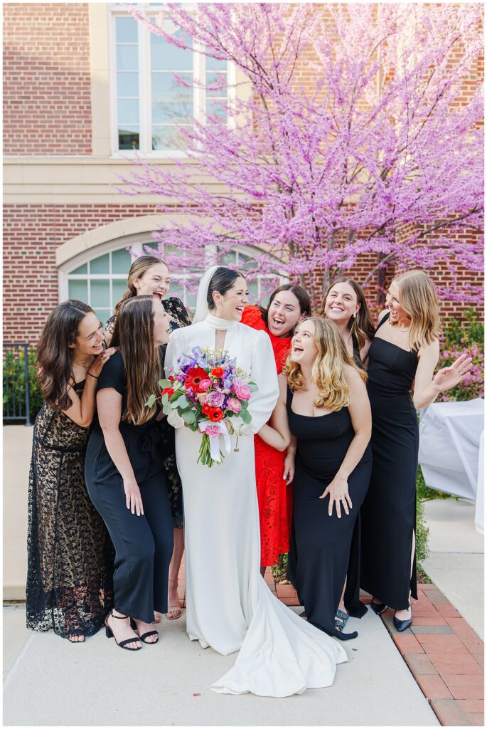 The bride poses outdoors with a joyful group of women during the wedding cocktail hour at the National Cathedral School, in front of a blooming pink tree and brick building; the bride holds a colorful bouquet and everyone is laughing.