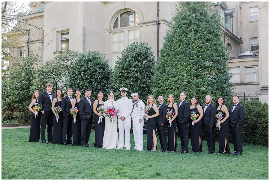 The full wedding party poses for a formal group portrait on the lawn during a National Cathedral School wedding. The bride and groom, both in white naval uniforms, stand at the center, surrounded by bridesmaids in black dresses holding colorful bouquets and groomsmen in black tuxedos. A stone building and neatly trimmed greenery form the background.
