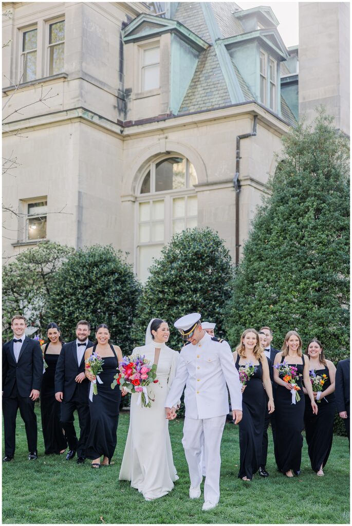 The bride and groom, both in white naval uniforms, walk hand-in-hand across the lawn with their wedding party at a National Cathedral School wedding. Bridesmaids in black dresses hold colorful bouquets, and groomsmen in black tuxedos walk alongside, all smiling in front of a stone building and manicured greenery.