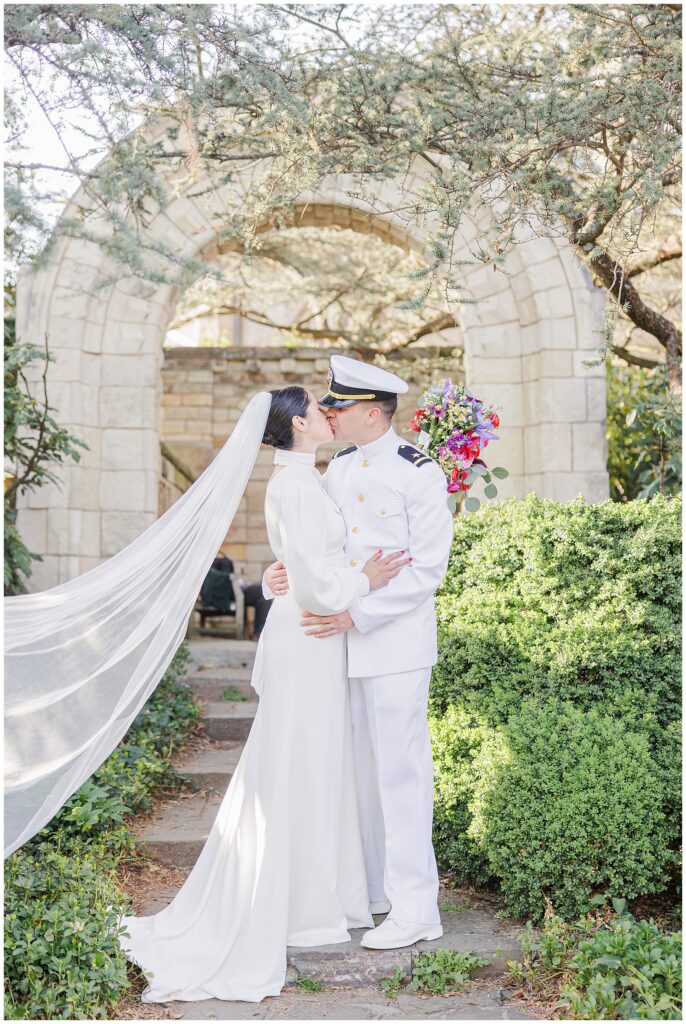 The bride and groom, in a white gown and white naval uniform, kiss beneath a stone archway surrounded by greenery in Bishop’s Garden at the Washington National Cathedral.