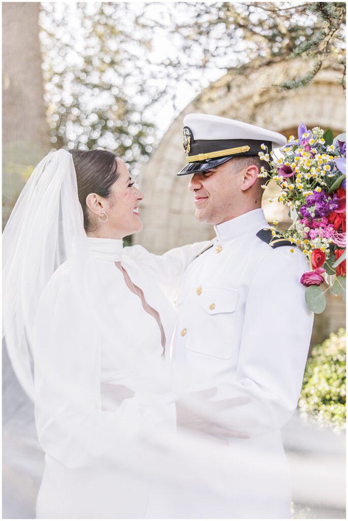 Close-up of the bride smiling at the groom while holding his shoulder, with the groom in a white naval uniform holding a bouquet, taken in Bishop’s Garden at the Washington National Cathedral.