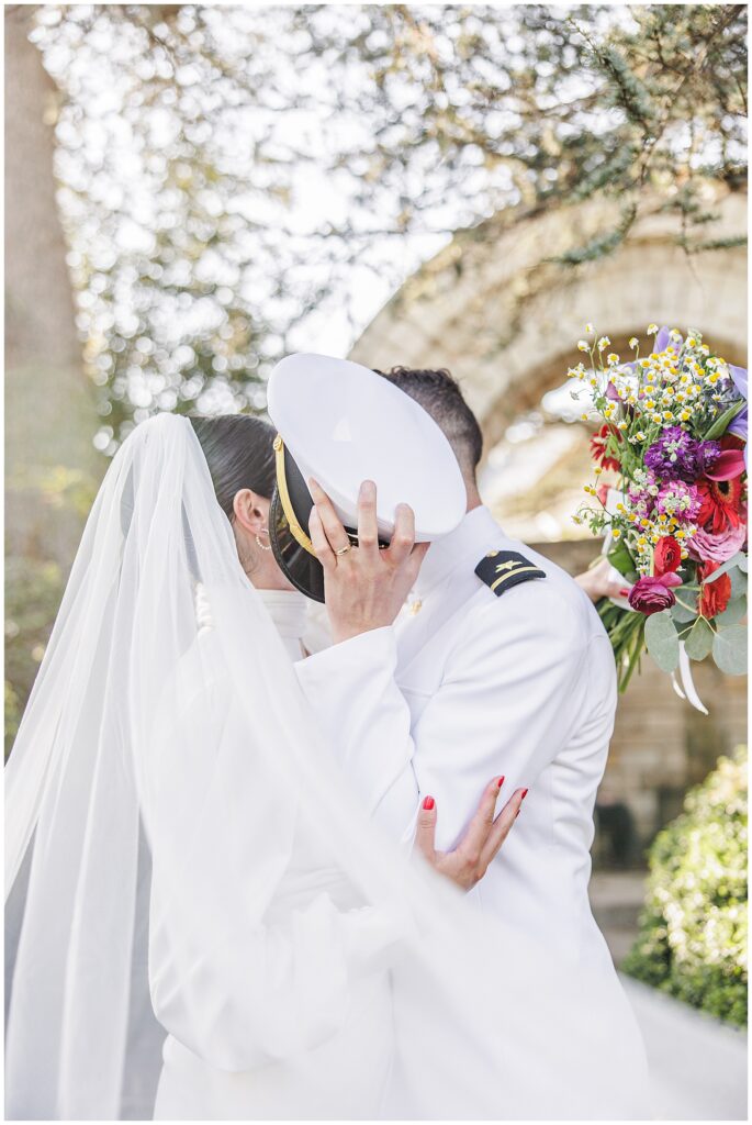 The bride and groom, in a white gown and white naval uniform, kiss beneath a stone archway surrounded by greenery in Bishop’s Garden at the Washington National Cathedral.