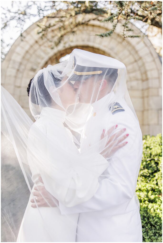 The bride and groom share a kiss beneath the bride’s flowing veil in Bishop’s Garden at the Washington National Cathedral, with a stone archway in the background.