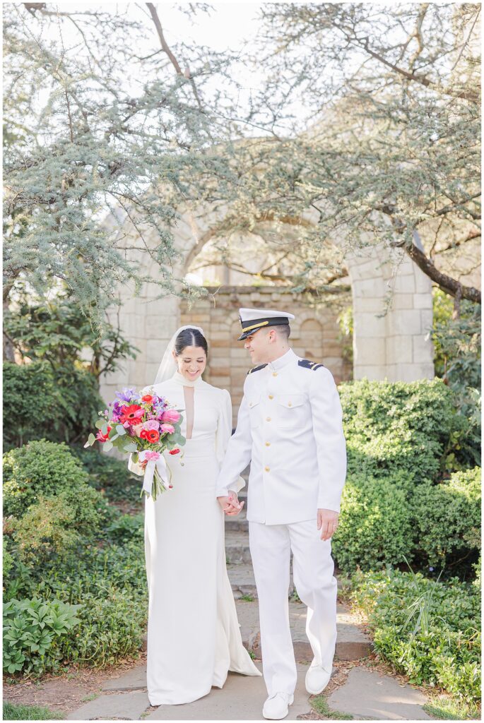 The bride and groom hold hands and smile while walking through Bishop’s Garden at the Washington National Cathedral, surrounded by lush greenery and a stone structure.