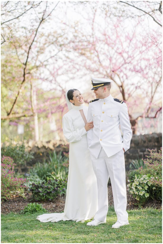 The bride and groom stand closely, smiling at each other in Bishop’s Garden at the Washington National Cathedral, with blooming cherry blossom trees in the background.
