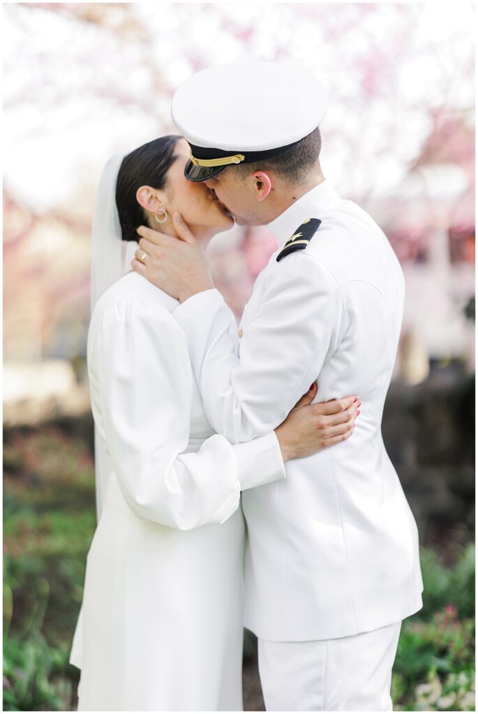 Close-up of the bride and groom kissing among spring blossoms in Bishop’s Garden at the Washington National Cathedral, both dressed in white wedding attire.