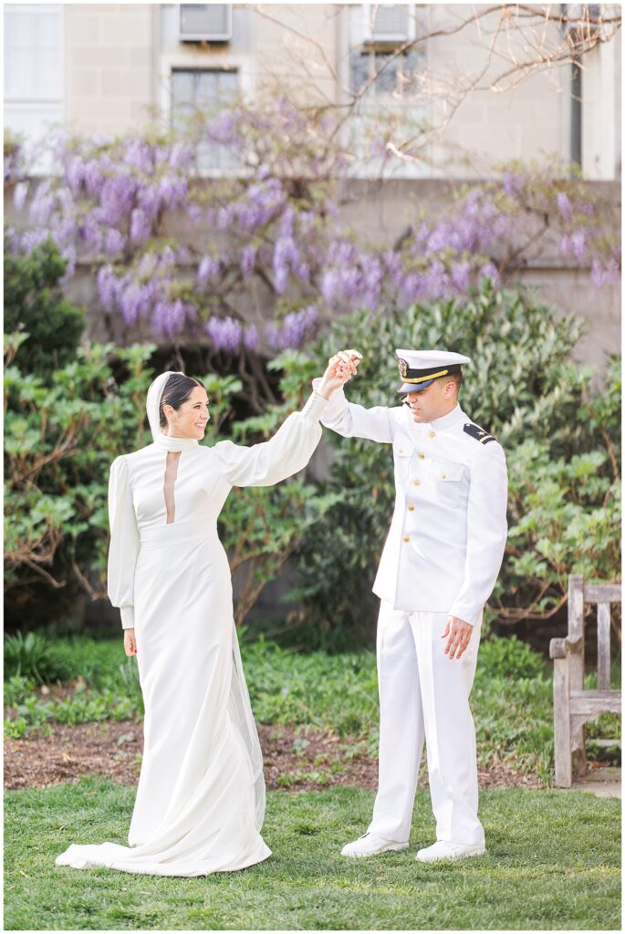 The groom in a white naval uniform twirls the bride, who wears a long-sleeved white gown and veil, as they dance on a lawn in Bishop’s Garden during a Washington National Cathedral wedding, with blooming purple wisteria in the background.