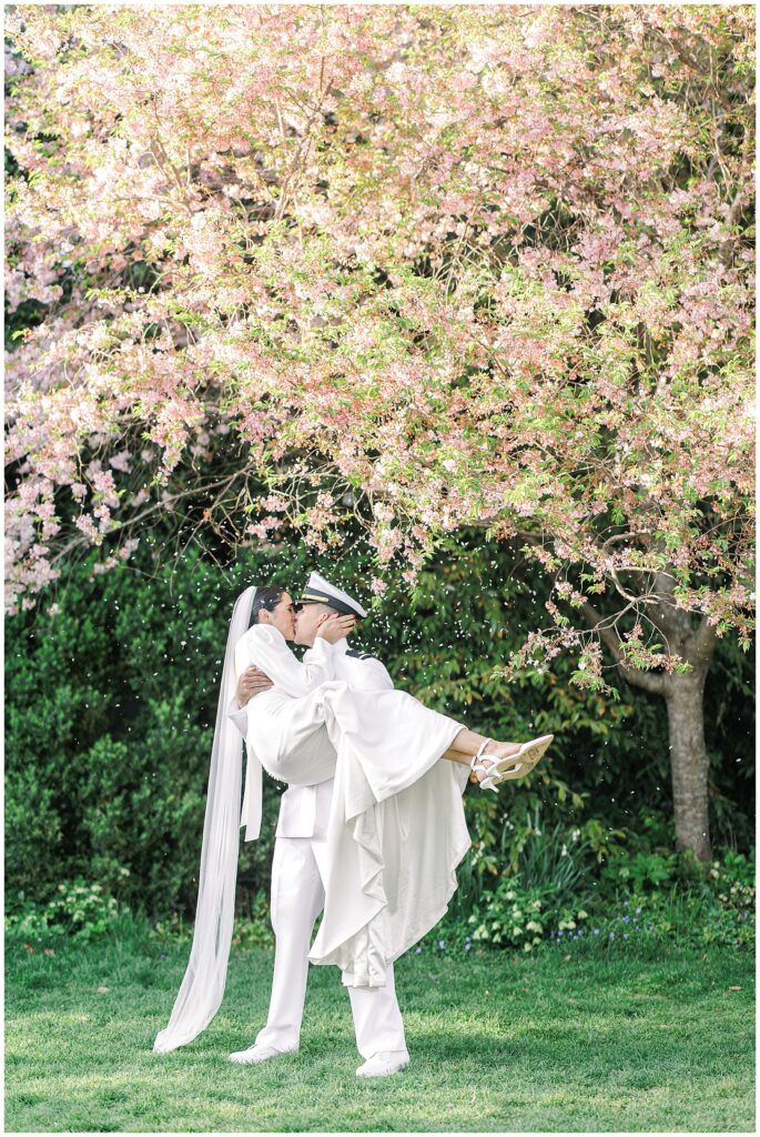 The groom lifts the bride in his arms and kisses her under a pink-blossomed tree in Bishop’s Garden during a Washington National Cathedral wedding, as petals fall around them.